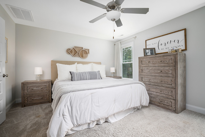 a bed with white sheets, nightstand, dresser with sign above it, carpet flooring