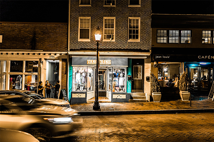 a street with a few shops at night, light in the middle, cars passing by 